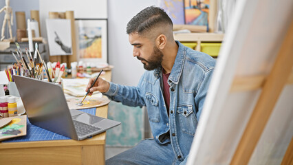 Handsome bearded man painting in an art studio, using a laptop for reference, showcasing creativity and study.