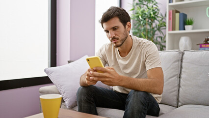 A handsome hispanic man focused on texting with his smartphone while sitting on a couch indoors.