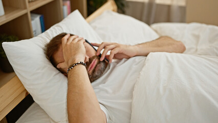A mature hispanic man with grey hair lying in bed, indoors, looking at his phone with a pensive expression.