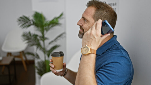 Mature hispanic man with grey hair talking on a phone indoors while holding a coffee cup, showcasing casual business style.