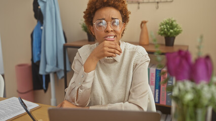 African american woman ponders while working from a cozy home office setup, portraying a casual, yet focused, remote work environment.