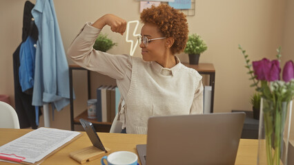 Confident african american woman flexing arm muscles in a cozy home office setting, portraying empowerment.
