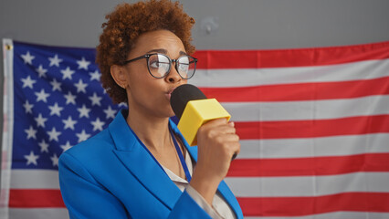 African american woman speaking into microphone with us flag backdrop in indoor setting.