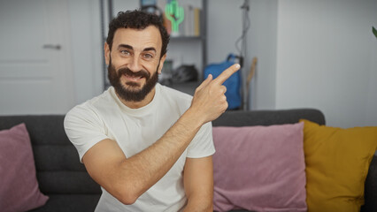 A cheerful bearded man in a modern living room pointing at something, displaying positivity and comfort.