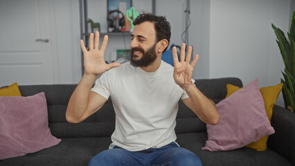 A smiling bearded man in casual clothing sitting on a grey sofa showing nine fingers indoors.
