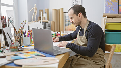 A focused man with a beard working on a laptop in a bright studio surrounded by art supplies.