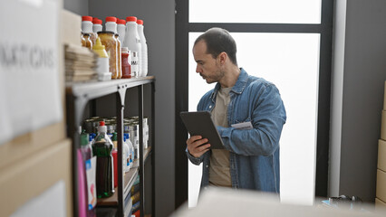 A focused man with a tablet volunteers at an indoor donation center, surrounded by shelves stocked with food and supplies.