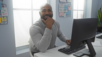 Fototapeta premium Handsome young african american man with a beard working at a computer in a bright office interior and smiling confidently at the camera