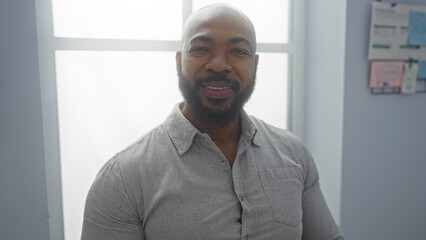 Young african american man with a beard and bald head smiling confidently in an office room with a window and notice board in the background.