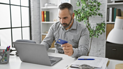 A mature bearded man with grey hair, holding a credit card, works on a laptop in a modern office.