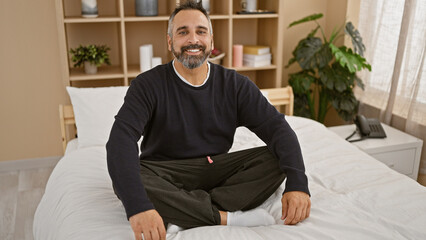 Fototapeta premium A mature hispanic man with grey beard smiling casually sitting on a white bed in a well-lit bedroom interior.