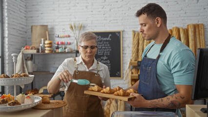 Woman and man working together in a bakery, assembling pastries on a tray, in an indoor shop setting full of baked goods and a chalkboard menu in the background.