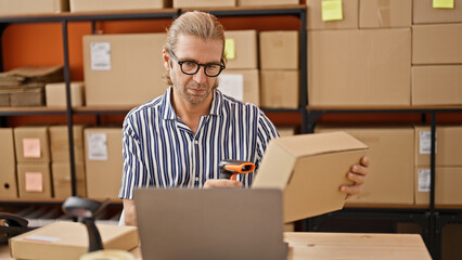 A middle-aged man works diligently in a warehouse, scanning a package amidst shelves filled with boxes.