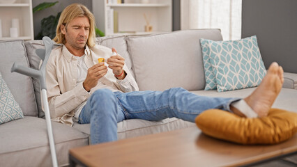 A blond man with long hair, analyzes medication while resting on a couch at home, with crutches nearby indicating injury or recovery.