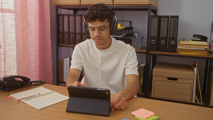 Young, hispanic man with headphones working on a tablet in an organized office, surrounded by...
