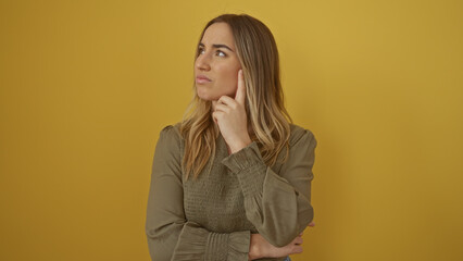A beautiful young blonde woman ponders thoughtfully with her finger on her cheek against a vivid...