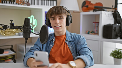 A youthful male podcaster with headphones interacts with his smartphone in a modern home studio setup.