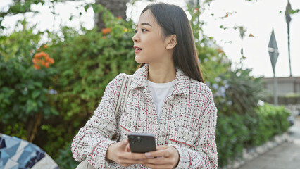 A young asian woman in a tweed jacket using a smartphone on a sunny green urban park pathway.