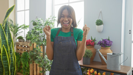 A cheerful african woman in a green shirt and denim apron celebrates inside a vibrant flower shop...