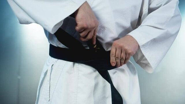 Karate fighter ties black belt at smoky background. Man stands confidently, proudly displaying black karate belt. Belt symbolizing skill and achievement in martial arts. Close-up in 4K, UHD
