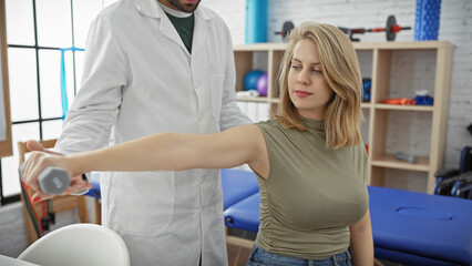 Obraz premium A woman exercises her arm with a dumbbell under a male physiotherapist's guidance in a well-equipped therapy room.