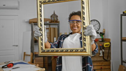 A smiling woman artist holding a picture frame in a creative carpentry studio with tools visible in...
