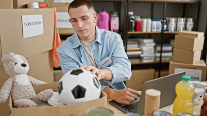 A focused young hispanic man volunteers at a donation center, sorting items, using a laptop against...