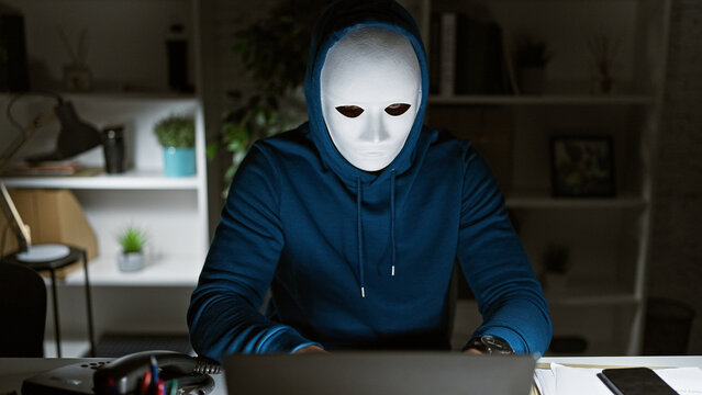 Mysterious person with a white mask and hoodie using computer in a dark office setting at night. - Powered by Adobe