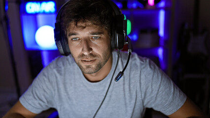 Handsome hispanic man with beard and green eyes wearing headphones in a dark gaming room at night