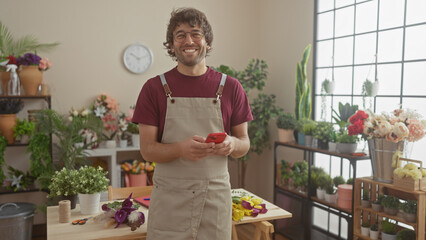 A cheerful man with glasses and beard in an apron holding a phone in a flower shop full of various plants.