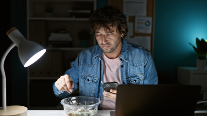 A young hispanic man in a denim jacket uses a smartphone while eating and working late in an office.