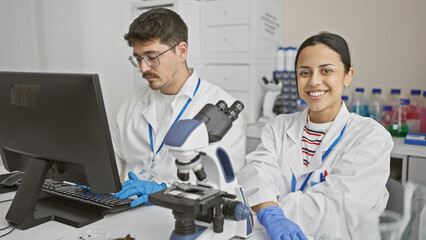 A man and woman, likely coworkers, are working together in a laboratory setting, surrounded by scientific equipment.