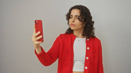 Naklejka premium Young hispanic woman taking a selfie with a red phone, isolated on a white background wearing a red jacket and white top.
