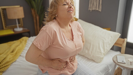 A mature blonde woman experiencing stomach pain sitting on a bed in a cozy bedroom at home.