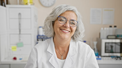 A smiling mature woman with grey hair wearing glasses and lab coat in a laboratory setting.