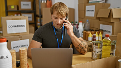 A young bearded man coordinates food donations in a warehouse while talking on the phone and using a laptop.