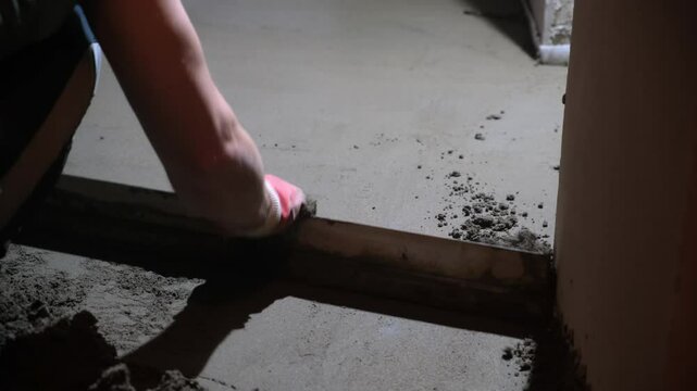 Builder worker making cement floor screed in rough finishing apartment. Repair master aligning fresh floor surface with spatula. Construction, overhaul, reconstruction work in unfinished building.