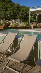 Two empty deck chairs sit by the poolside on a sunny day, surrounded by lush greenery and a patio area with wooden chairs in the background.