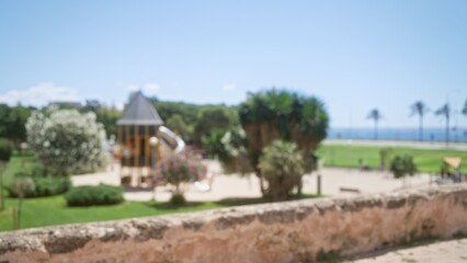 Obraz premium Blurred sunny mallorca outdoor scene with playground surrounded by lush greenery and defocused trees under a clear blue sky
