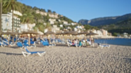 Blurred beach scene in mallorca with people enjoying a sunny day on lounge chairs under straw umbrellas, mountains and buildings in the background.