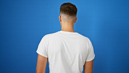 Back view of a young adult hispanic man isolated against a blue background, wearing a white t-shirt.