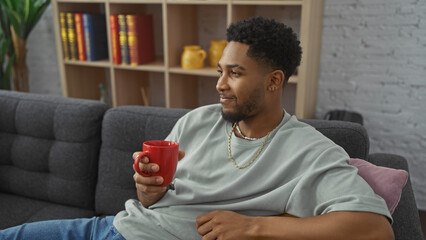 A relaxed african american man holding a red mug while sitting comfortably on a grey couch indoors with a bookshelf background.