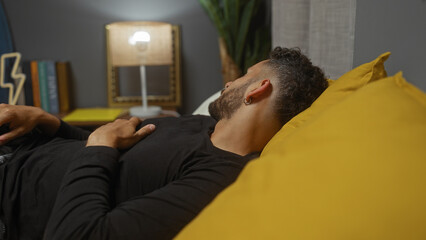 Fototapeta premium A young hispanic man lying comfortably in bed within a cozy and well-decorated bedroom in his home.