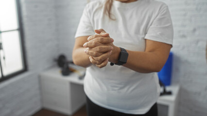 Elderly hispanic woman at indoor gym with hands clasped together wearing white shirt and smartwatch, showing a close-up view focusing on her hands.