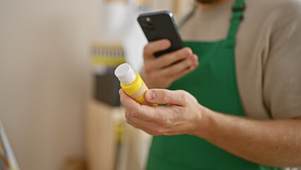 A young bearded man in an apron scans a product with his smartphone in a bright room.