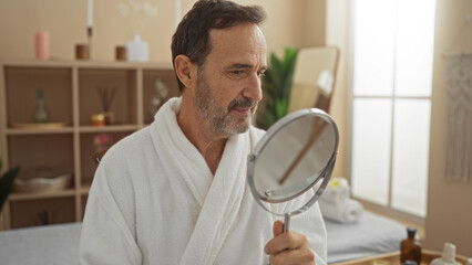 Mature hispanic man in a spa wearing a white robe, holding a hand mirror, and checking his face in a serene wellness room.