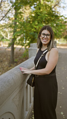Smiling young woman with glasses dressed in black enjoying a sunny day in a leafy park.