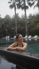 Fototapeta premium Smiling young woman relaxing in a luxurious bali resort swimming pool surrounded by tropical palms.