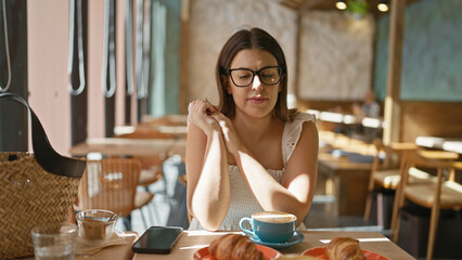 Beautiful hispanic woman enjoying her hot coffee drink inside sunny cafe, relaxing at breakfast while wearing glasses, sitting casually at cafeteria table enjoying the morning sun.