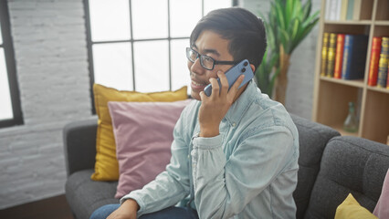 A young asian man sits comfortably indoors, smiling while talking on a cellphone in a modern living room.